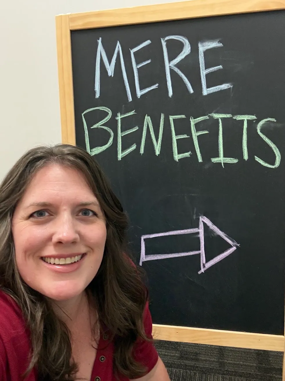 Kate Spilsbury smiling in front of a chalkboard that reads Mere Benefits with an arrow